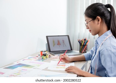 Beautiful young Asian fashion tailor with textile sewing accessories and entrepreneur designer sketches are full of bright colors on the desk with a laptop for creative ideas in the studio.