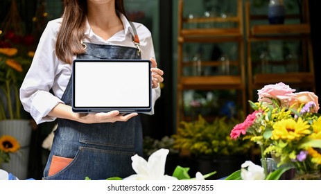 Beautiful Young Asian Female Florist Or Floral Shop Owner In Apron Showing Or Holding A Digital Tablet White Screen Mockup, Standing In Front Of Her Flower Shop. Cropped Shot