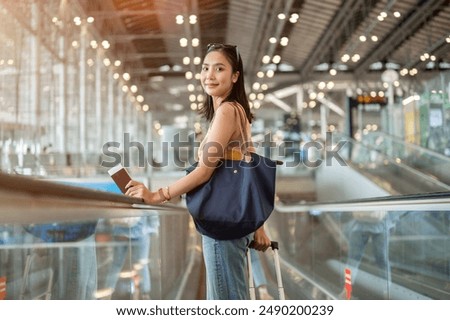 A beautiful young Asian female tourist passenger with her luggage is on the airport escalator, smiling at the camera, traveling by plane. people and transportation concepts, with copy space