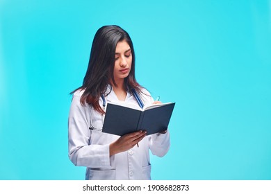 Beautiful Young Asian Girl Doctor, With A Notebook For Records Isolated On A Blue Background. Medical Student General Practitioner. The Concept Of Medical Education In India