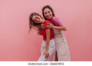 Beautiful young asian girl hugs her caucasian girlfriend after long absence indoors. Blonde glasses in red T-shirt and white pants along with brunette denim dress. Summer playful mood concept