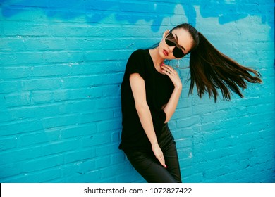 Beautiful young asian girl with long hair wearing sunglasses and black t-shirt posing in front of blue wall