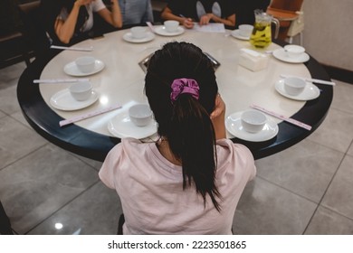 Beautiful Young Asian Girl With Ponytail And Satin Scrunchie Hair Tie Seated In Front Of A Chinese Round Table With Empty White Plates And Group Of People