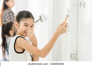 Beautiful Young Asian Kid Girl Model In Painting White Wall And Posing Closing Nose With Her Hands For Bad Smell, Painting The Wall And Looking At Camera. Studio Shot.