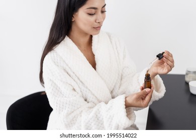 Beautiful young asian model holding opened skincare product in glass bottle with pipette while sitting at makeup table in white bathrobe at home. Natural organic cosmetic and skincare concept