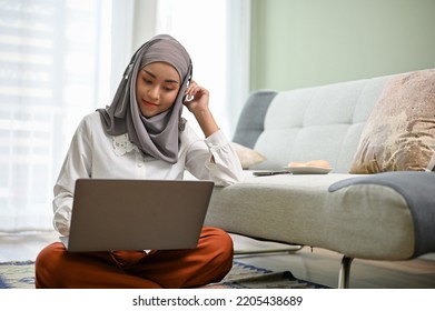 Beautiful Young Asian Muslim Woman Wearing Hijab Using Laptop Computer, Planning Her Project And Sitting In Her Living Room. Work From Home Concept