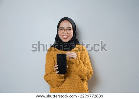 Beautiful young asian muslim woman wearing orange sweater and glasses pointing phone at camera over isolated background
