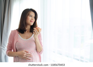 Beautiful Young Asian Pregnant Woman Holding Red Apple By Left Hand Standing In Front Of Window. Her Right Hand Is Touching Her Tummy With Love Expression. Modern Healthy Mom Concept.