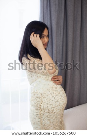 Beautiful young Asian pregnant woman standing by the window and touching her tummy. Pregnancy. Health. Motherhood. A cozy portrait of a pregnant woman.