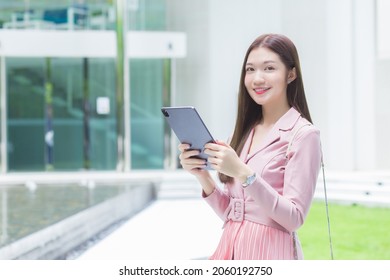 Beautiful Young Asian Professional Business Woman With Long Hair Is Smiling Outdoor In The Garden Front Of The Office While Hold At The Tablet In Her Hand To Go Out Meeting With Clients At The Office.