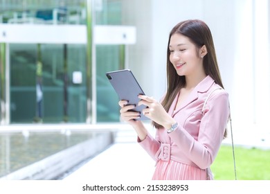 Beautiful Young Asian Professional Business Woman With Long Hair Is Smiling Outdoor In The Garden Front Of The Office While Hold At The Tablet In Her Hand To Go Out Meeting With Clients At The Office.