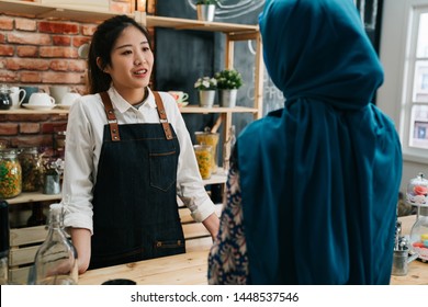 Beautiful Young Asian Restaurant Female Owner In Apron Talking With Muslim Customer In Blue Hijab At Counter In Morning Coffee Shop. Lady Waitress Taking Order With Malay Woman In Modern Cafe Bar.