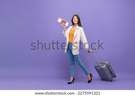 Beautiful young Asian tourist woman walking with trolley suitcase, passport and air ticket about to fly, studio shot purple color isolated background