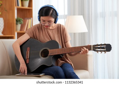 Beautiful young Asian woman with acoustic guitar sitting on sofa and concentrating on learning to play using app on tablet placed nearby  - Powered by Shutterstock