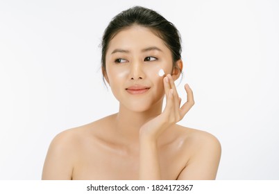 Beautiful young Asian woman with bare shoulders is applying face cream on her face and and looking away isolated over white background.Woman beauty face skin care concept.
