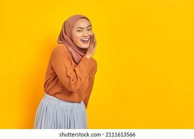 Beautiful young Asian woman in brown sweater and hijab hand on mouth telling secret, whispering gossip, looking at camera with cheerful expression on yellow background. People islam religious concept - Shutterstock ID 2111613566