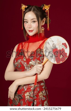 Beautiful young Asian woman with clean fresh skin wearing traditional cheongsam qipao dress holding fan posing on red background. Portrait of female model in studio. Happy Chinese new year.