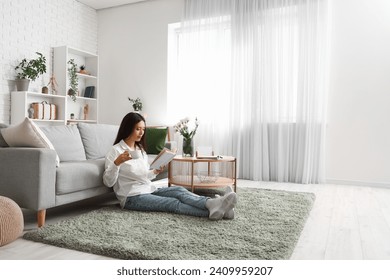 Beautiful young Asian woman with cup of tea reading book in living room - Powered by Shutterstock