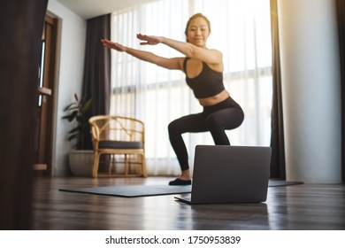 A beautiful young asian woman doing squats while watching online workout tutorials on laptop at home