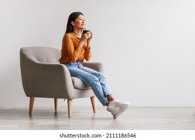 Beautiful Young Asian Woman Drinking Coffee Or Tea, Having Break, Relaxing In Armchair Against White Studio Wall, Copy Space. Millennial Female Chilling And Enjoying Hot Beverage