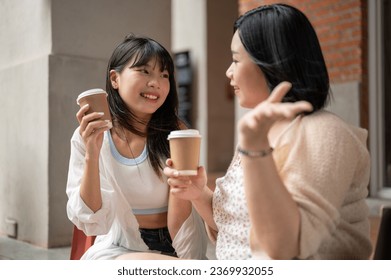 A beautiful young Asian woman enjoys talking and having coffee with her friend while sitting on the stairs in the city. Urban lifestyle concept - Powered by Shutterstock