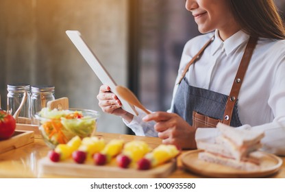 A Beautiful Young Asian Woman Following Recipe On Digital Tablet While Cooking Salad And Sandwich In The Kitchen, Online Learning Cooking Class Concept