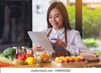 A Beautiful Young Asian Woman Following Recipe On Digital Tablet While Cooking Salad And Sandwich In The Kitchen, Online Learning Cooking Class Concept