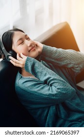 Beautiful Young Asian Woman With Headphones Relaxing On The Sofa. She Is Listening To Music Using Smartphone . Chill Out And Leisure Concept
