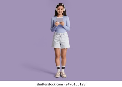 Beautiful young Asian woman in headphones listening to audiobook with modern tablet computer on lilac background - Powered by Shutterstock