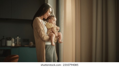 Beautiful Young Asian Woman Holding her Baby in her Arms While Standing Next to a Window at Home. Cute Little Toddler Resting in His Mother's Embrace as She Shows him the Busy Streets of The City. - Powered by Shutterstock