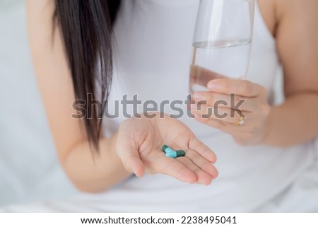 Beautiful young asian woman holding drug and glass of water while ill sitting on bed in the bedroom at home, female fever and healthcare while uncomfortable and unhappy, medical and lifestyle concept.