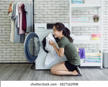 Beautiful Young Asian Woman Housewife Sitting With Smiling And Smelling White Clean Towel After Washing From Washing Machine In Laundry Room.