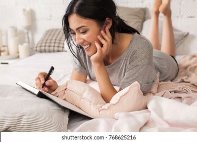 Beautiful young asian woman laying on bed and writing a diary. Smiling brunette lady with notebook and pen in her hands. Modern bright home interior on background.