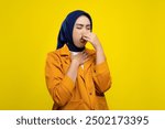 Beautiful young Asian woman pinching her nose with fingers, smelling something smelly and disgusting isolated on yellow background