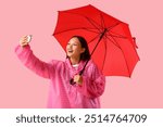 Beautiful young Asian woman in raincoat with umbrella taking selfie on pink background