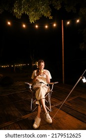 Beautiful Young Asian Woman Relaxing Sipping A Hot Cocoa Or Coffee, Sitting On A Camping Chair In The Beautiful Camping Site At Night.