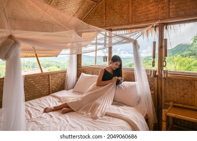 Beautiful Young Asian Woman Resting And Looking At Retro Camera On The Bed In Wooden Cottage Among The Mountain On Vacation