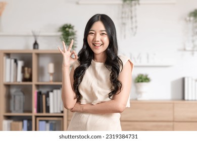 Beautiful young Asian woman showing thumbs up OK gesture smile with positive emotional at home. Attractive female making okay hand sign or say yes happiness and cheerful. Women lifestyle concept