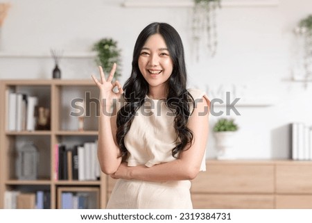 Beautiful young Asian woman showing thumbs up OK gesture smile with positive emotional at home. Attractive female making okay hand sign or say yes happiness and cheerful. Women lifestyle concept