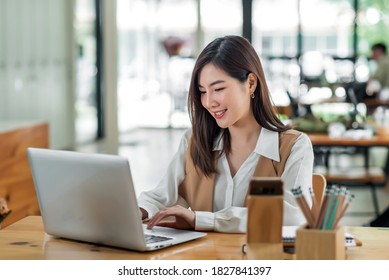 Beautiful Young Asian Woman Sitting At Coffee Shop Using Laptop. Happy Young Businesswoman Sitting At Table In Cafe With Tab Top Computer.