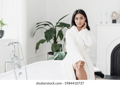 Beautiful Young Asian Woman Sitting In Light Spacious Bathroom With Green Plants Wearing White Bathrobe Preparing For Pampering Morning Routine. Beauty And Selfcare Concept