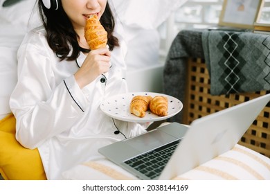 Beautiful Young Asian Woman Sitting Next To Her Bed Using Her Laptop And Enjoy Breakfast In The Morning At Home. Small Business Owners Are Checking Orders Online. Concept For Freelancer Lifestyle.