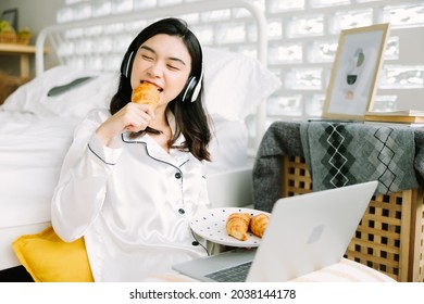 Beautiful Young Asian Woman Sitting Next To Her Bed Using Her Laptop And Enjoy Breakfast In The Morning At Home. Small Business Owners Are Checking Orders Online. Concept For Freelancer Lifestyle.