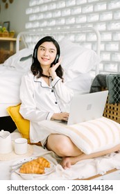 Beautiful Young Asian Woman Sitting Next To Her Bed Using Her Laptop And Enjoy Breakfast In The Morning At Home. Small Business Owners Are Checking Orders Online. Concept For Freelancer Lifestyle.