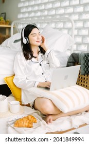 Beautiful Young Asian Woman Sitting Next To Her Bed Using Her Laptop And Enjoy Breakfast In The Morning At Home. Small Business Owners Are Checking Orders Online. Concept For Freelancer Lifestyle.