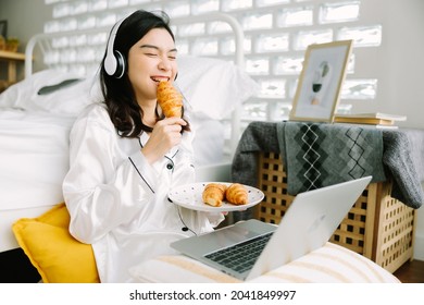 Beautiful Young Asian Woman Sitting Next To Her Bed Using Her Laptop And Enjoy Breakfast In The Morning At Home. Small Business Owners Are Checking Orders Online. Concept For Freelancer Lifestyle.