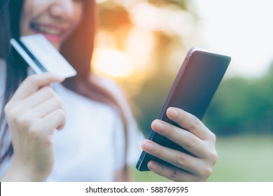 Beautiful Young Asian Woman Smiling While Using Her Smartphone To Shop Online With Credit Card In Garden Background. Portrait Of Asian Women Shopping By Creditcard With Smart Phone In Park Outdoor. 