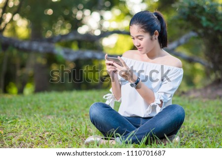 Beautiful Young asian woman smiling while reading her smartphone in garden park outdoor. Girl hand holding using mobile phone concept.
