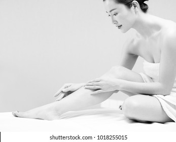 Beautiful Young Asian Woman With Soft Skin Wrapped In White Towel Sitting On Bed Applying Body Lotion, Black And White.