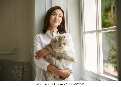 Beautiful Young Asian Woman Standing By Window At Home Holding A Cat In Arms Looking Serene And Content